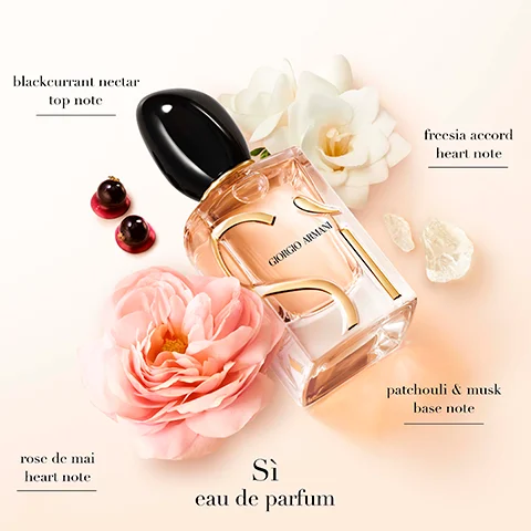 A Giorgio Armani Sì eau de parfum bottle with a black cap and gold detailing is presented on a light background surrounded by a pink rose, white flowers, two dark berries, and light colored crystals, with visible text reading blackcurrant nectar top note, freesia accord heart note, patchouli & musk base note, rose de mai heart note, Sì eau de parfum, and GIORGIO ARMANI.